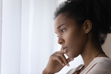 Close up head shot side view young african american thoughtful woman standing near window, looking...
