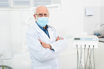 senior dentist in white coat and medical mask standing with crossed arms in dental clinic.
