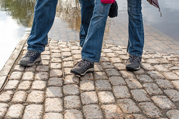 Man and woman tourists in blue jeans and comfortable sneakers stand on paved road flooded by river on spring day close view