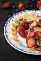 Some pancakes with sweet strawberry, blueberry and banana sliced on black table with some fruit on it 