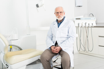 senior dentist in white coat sitting with clenched hands in dental clinic.