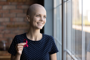 Happy hairless girl with oncology disease holding red ribbon, showing symbol of fight against...