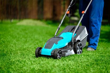Close-up of a man in overalls with a lawn mower cutting green grass in a modern garden. Lawn mowing...