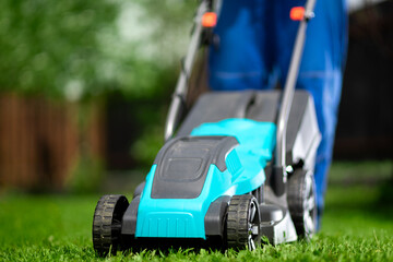 Close-up of a man in overalls with a lawn mower cutting green grass in a modern garden. Lawn mowing machine. - obrazy, fototapety, plakaty