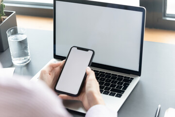 Close up businesswoman using smartphone and laptop with empty white blank screen mockup, sitting at desk, employee worker consulting client online, preparing presentation, browsing mobile device apps