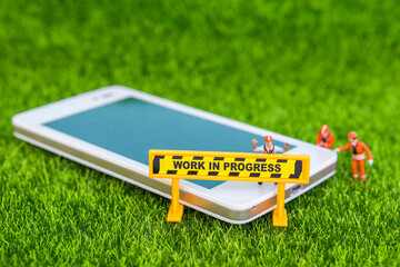 Work in progress sign with miniature worker and smartphone on green grass background, technology business, phone repair service 