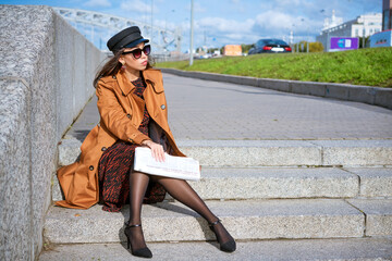 Young woman in sunglasses of Caucasian ethnicity sits on the steps on the embankment in a black cap and brown jacket with a newspaper in her hand