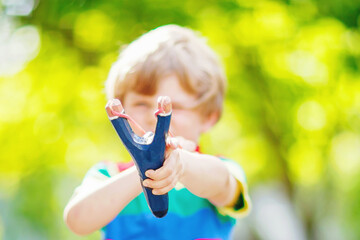 Funny little kid boy shooting wooden slingshot against green tree background. Child having fun in...