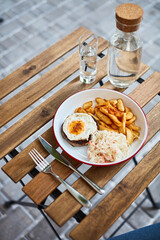 schnitzel with egg, potatoes and cabbage on a wooden table on the street terrace summer with a fork and knife with a glass jug of water