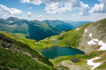 Fototapeta na wymiar capra lake of fagaras mountains. wonderful summer nature scenery on a sunny day. popular travel destination of romania. snow and grass on the slopes. fluffy clouds on the sky