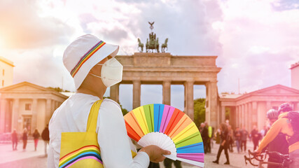 Mature Caucasian LGBT woman in Berlin with rainbow fan and ribbon on white summer hat. Rainbow, symbol of LGBTQIA gay lesbian pride and diversity including covid dissidents. Woman wearing PP2 mask.