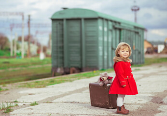 charming baby in a red coat with a suitcase is waiting for a train on the platform