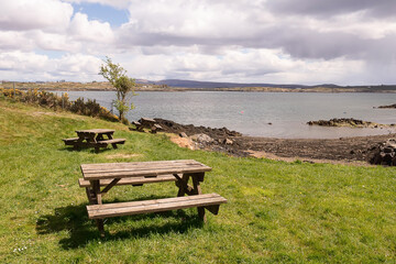 Naklejka na ściany i meble Picnic area by the ocean with beautiful view. Wooden benches and tables on the grass. Warm sunny day. Roundstone town, county Galway, Ireland. Nobody.