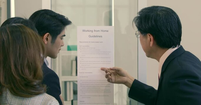 Asian boss explain work from home guidelines to his colleague by pointing and showing at piece of paper attached on the glass wall in office. Concept of new normal