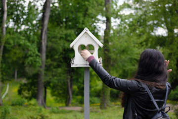 girl feeds birds in a feeder in an spring park, back view. girl puts food. taking care of birds, natural green background, bokeh, focus. wooden birdhouse. white bird feeder and woman in the garden