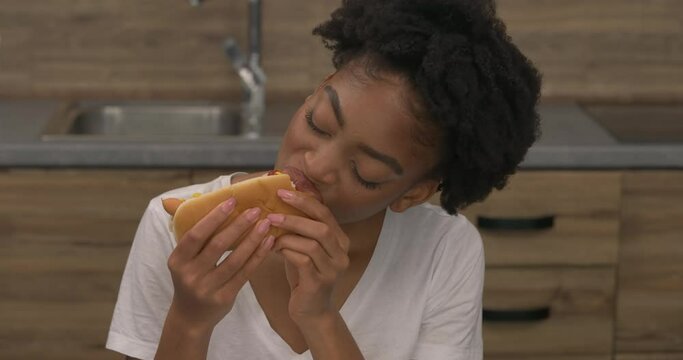 Tilting camera of African woman eating hot dog at home. Fast food. Junk food