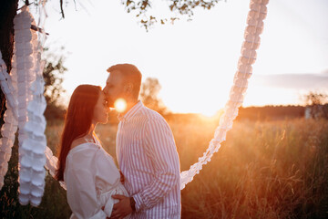 the sun brightly illuminates the couple in love with orange light. young husband kisses his pregnant wife on the forehead in the field. a place in the park decorated with white paper garlands for phot