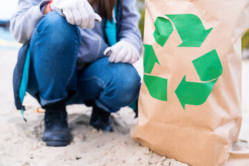 Female volunteer is collecting and sorting garbage on ocean river beach, folding waste trash in eco bag with recycle logo. Girl puts effort into making planet better. Contamination of nature.