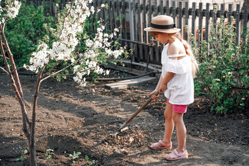 A farmer girl in a hat in the garden is engaged in gardening, a rake for harvesting leaves in her...