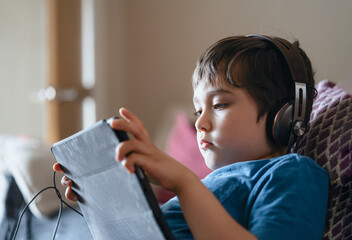 Child lying on couch wearing headphone listening to music, Close up face Kid sitting on sofa watching cartoons on tablet,Young boy playing game on touch pad or relaxing on his own in living room