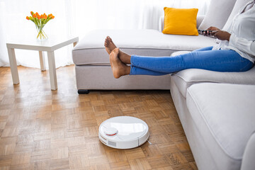 Robot vacuum cleaner cleaning carpet, woman legs rest sitting on sofa at home. Woman lifts feet up...