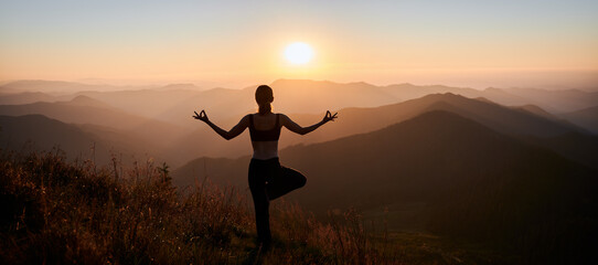 Back view of woman practicing yoga on background of evening mountains. Meditating female is balancing on one leg after sunset. Panorama.