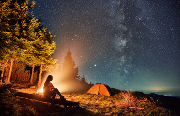 Young woman hiker sitting on bench near bonfire under magical sky with stars and Milky way....