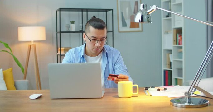 Young asian man shopping online sitting in designeted home office on laptop using credit card. Employee making payment transaction from home on digital notebook conected to internet