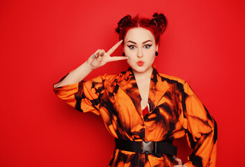 Beautiful girl on a red background. Red hair and tunnels in the ears, red dress and lips. Anime poses. The girl with eyeliner and two buns on her head grimaces, smiles, has fun. Peace gesture
