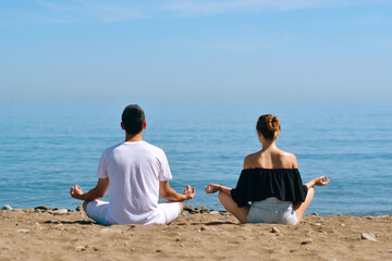 A couple is engaged in yoga on the beach against the background of the sea. Health and sports. A woman and a man on the shore of the ocean are meditating and relaxing. Summer and travel.