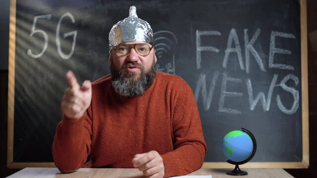 A bearded Caucasian male video blogger is broadcasting in his studio. A foil hat is worn on the head. Inadequate psycho talks about conspiracy theory.