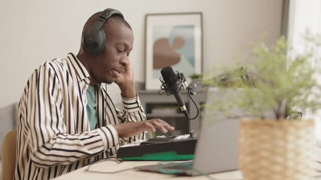 Medium shot of young African-American man wearing headphones sitting at desktop at home and singing in microphone while making electronic music using laptop and synthesizerMedium shot of young African