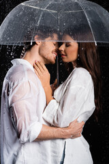 tender young adult couple standing in rain with umbrella and kissing on black background