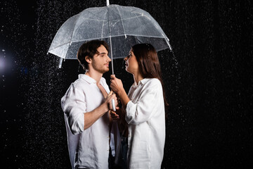 young adult boyfriend and girlfriend standing in rain with umbrella on black background