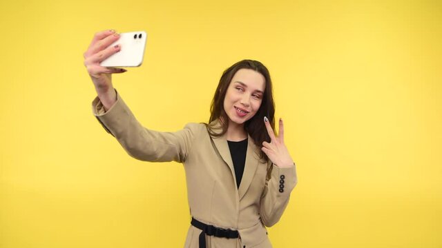Joyful lady in a jacket stands on a yellow background with a smile on her face takes a selfie and shows a gesture of peace, posing in the smartphone camera.
