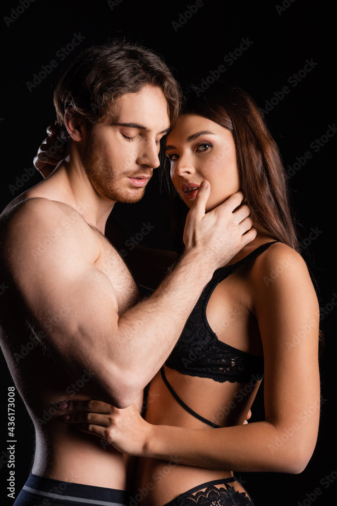 Wall mural muscular man with shirtless torso and closed eyes gently touching woman in lingerie isolated on blac