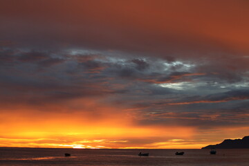 Fiery sunset landscape at sea over a lagoon with fishing boats. The silhouette of an island on the horizon with an orange sky and a large shiny metallic silver cloud.Hot image of a tropical evening