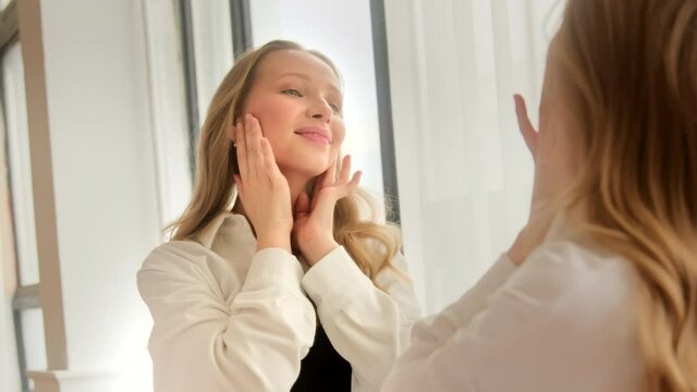 Young blonde woman looking touching face in the mirror examining skin condition and enjoy her healthy skin. Beauty product concept.