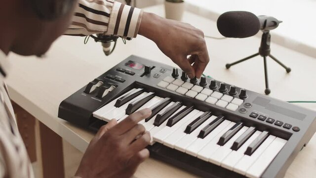 Back view of unrecognizable African-American man playing midi keyboard synthesizer while creating music in home studio