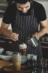 Young Asian barista man on black-and-white apron using spoon to deliberately topping coffee with milk froth for softness and mellowness at counter surrounding by dishes, jars, glass