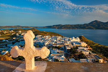 Christian cross and Plaka village on Milos island over red geranium flowers on sunset in Greece