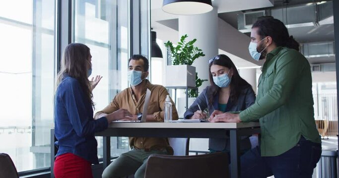 Diverse group of business colleagues wearing face masks sitting at table and talking