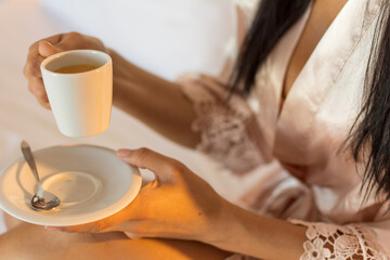 A young female, wearing a skin-colored nightdress and holding a cup of coffee in her hands in bed on a white blanket. Concept of a relaxed morning in bed.