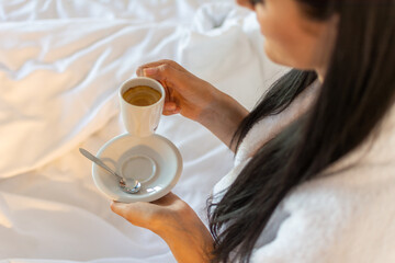 A young female, wearing a bathrobe, holding a cup of coffee in her hands in bed on a white blanket....