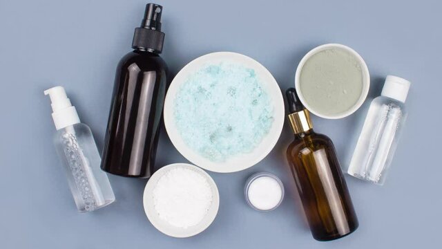 Moving picture of sea salt, serum in brown dropper glass and spray bottle, clay mask, jar of cream flat lay on blue background top view. Organic, natural cosmetic. Beauty, skincare, spa product