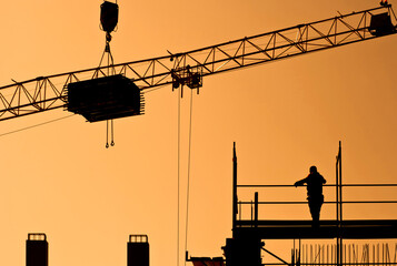 Worker on scaffolding and load hanging on crane at construction site, silhouettes in sunset