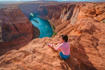 A beautiful young woman sits on the edge of a cliff overlooking Horseshoe Bend, the horseshoe...