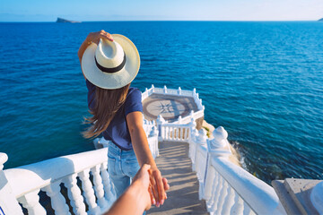 Young carefree tourist woman in white hat leading her boyfriend to the sea view. Follow me. Couple...