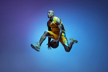 Sportive athletic african-american male basketball player training in neon light on blue background.