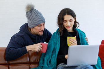 Caucasian young bearded husband in jacket knitted hat and pretty wife cover by blanket sit on couch drink hot coffee when browsing surfing internet from laptop computer together in winter at home